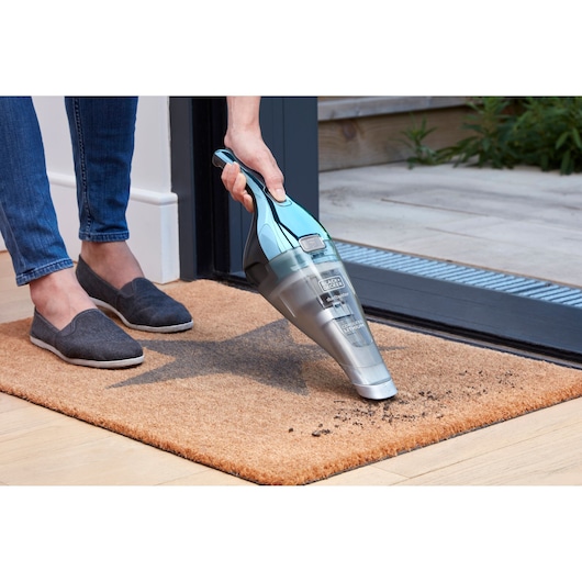 A person is using a BLACK+DECKER handheld vacuum cleaner to remove dirt from a brown doormat placed at the entrance of a house.