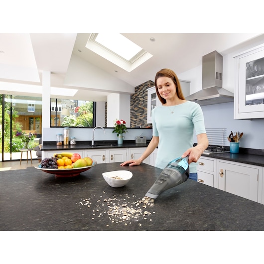 A person cleaning spilled cereal on a kitchen countertop with a BLACK+DECKER handheld vacuum cleaner model NVC220WBC. The kitchen has modern decor with fruit and a bowl on the counter.