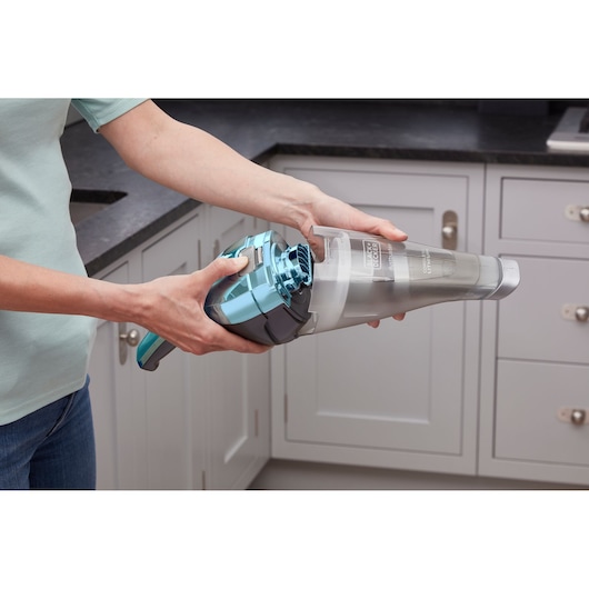 A person is holding a BLACK+DECKER handheld vacuum cleaner, showing the dust container being detached in a modern kitchen setting.