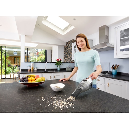 A person using a BLACK+DECKER handheld vacuum cleaner to clean spilled oats from a kitchen countertop. There is a bowl and a plate of assorted fruit on the counter, and the kitchen is bright and modern with large windows.