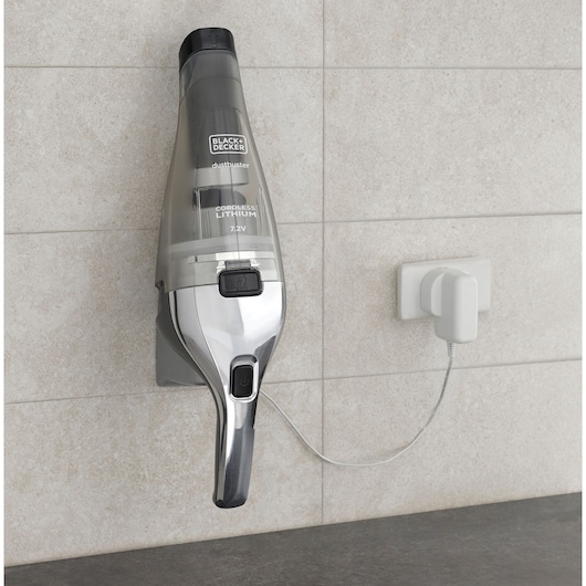 A BLACK+DECKER cordless handheld vacuum cleaner mounted on a charging station attached to a tiled wall, with the charging cable plugged into a wall socket.