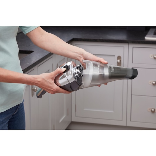A person is holding and opening a BLACK+DECKER handheld vacuum cleaner in a kitchen setting.
