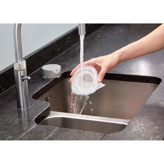 A person rinsing a removable vacuum filter under running water at a kitchen sink.