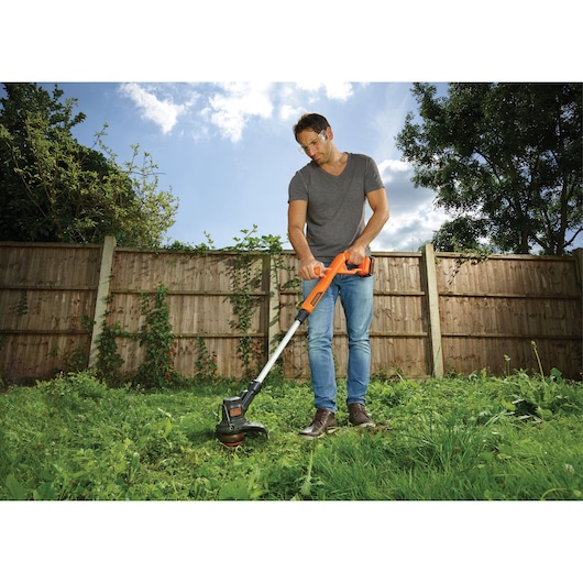 BLACKDECKER ST182320 orange cordless grass trimmer used in a garden, showing trimming action near a fence.