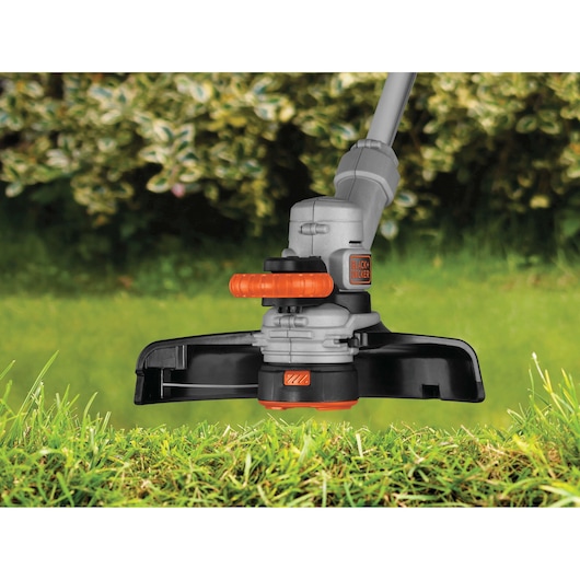 Photo of BLACKDECKER STC1820 grass trimmer head in action on green lawn, showing orange handle and safety guard.