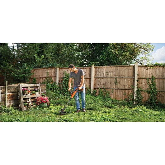 BLACKDECKER STC1840EPC cordless grass trimmer in use in a garden with wooden fence and plants.