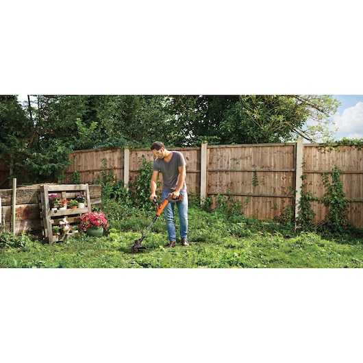 A person using a BLACK+DECKER cordless grass trimmer in a garden with wooden fencing, surrounded by plants and flowers. The person's face is blurred.